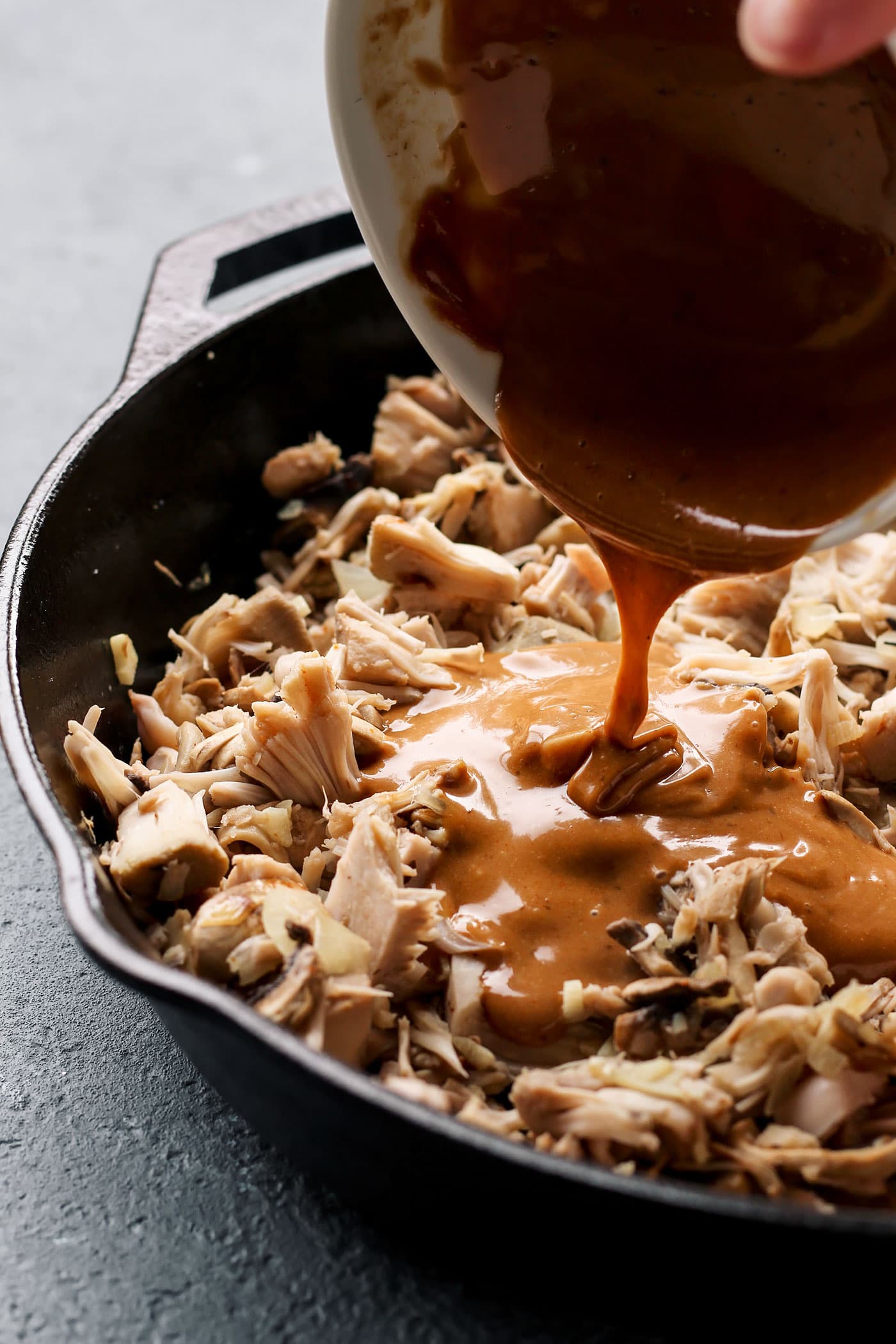 Pouring satay sauce into a skillet containing shredded jackfruit.