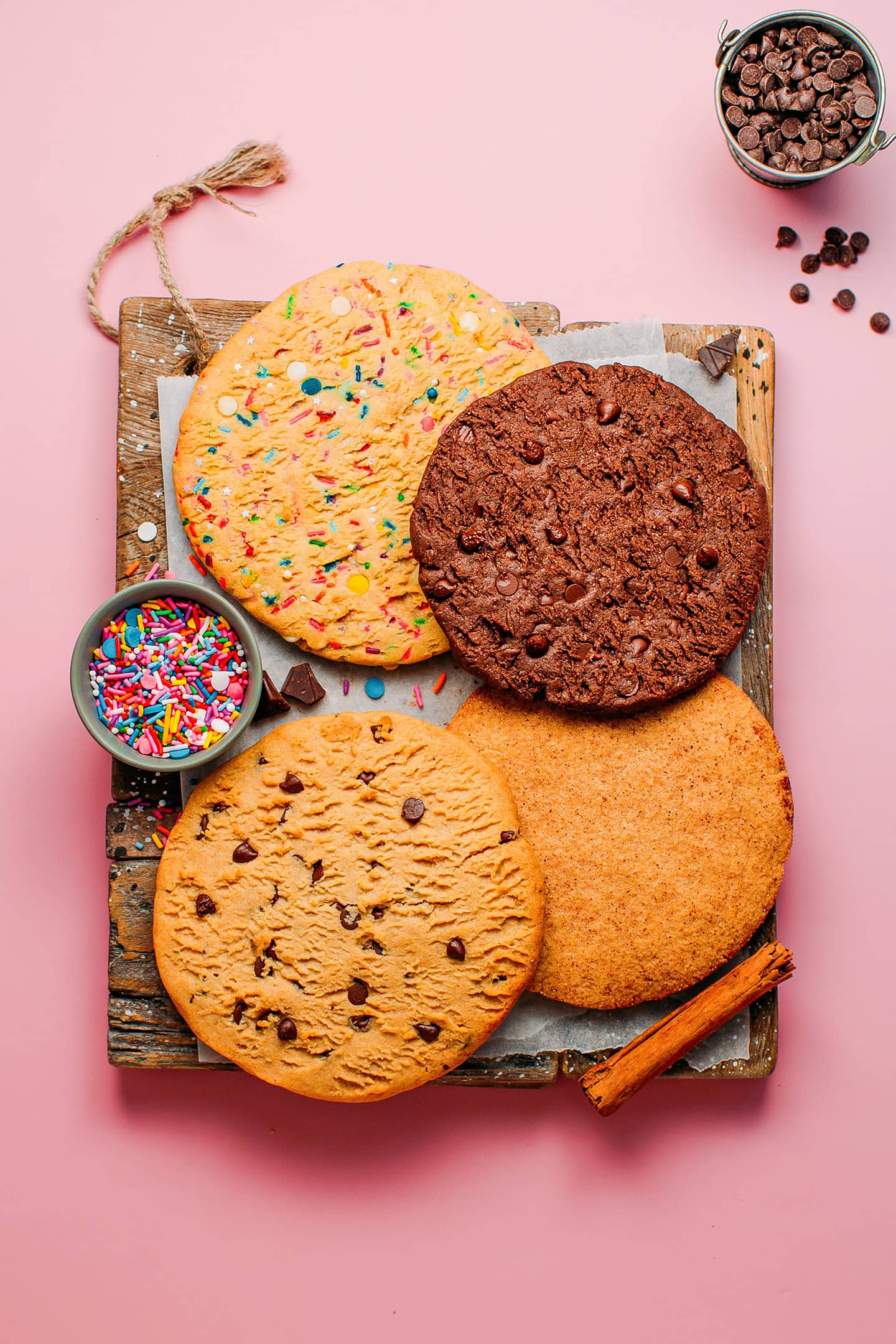 Four different cookies with chocolate chips, sprinkles, cinnamon sugar, and chocolate, on a wooden board.