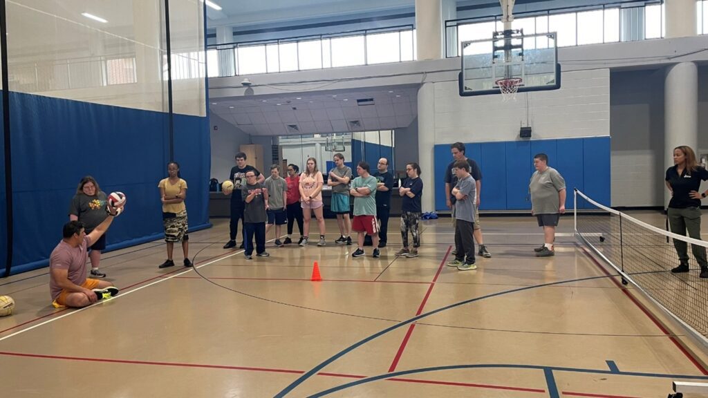 A group of individuals in a gymnasium watching as they receive a demonstration about how to play sitting volleyball.