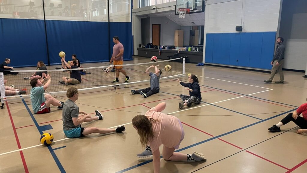 Many people seated on a gymnasium floor hitting volleyballs back and forth to each other. There is an adapted volleyball net between them.