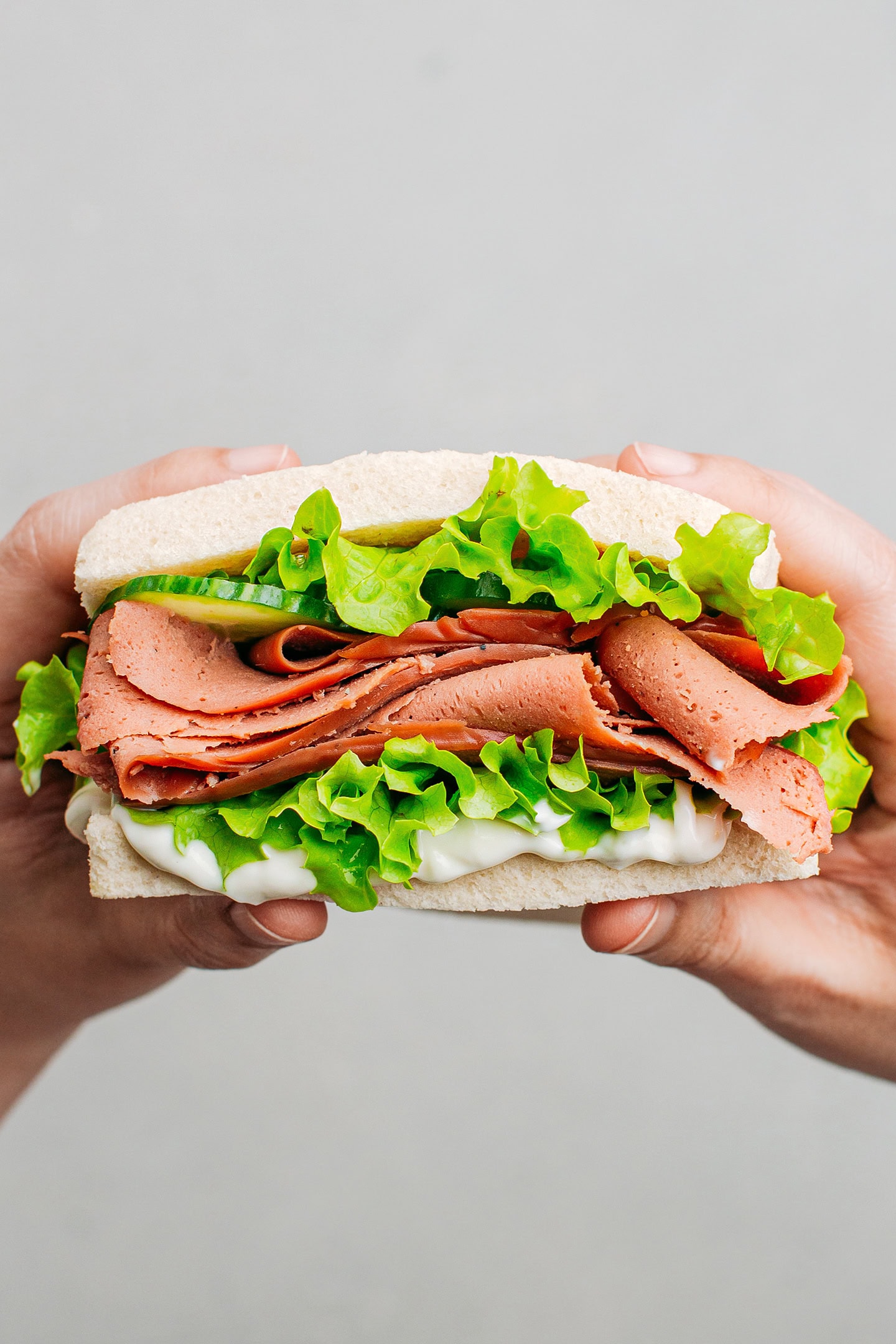 Holding a sandwich filled with vegan deli slices, mayo, cucumber, and lettuce.
