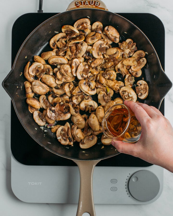 Pouring cognac into a skillet filled with sautéed mushrooms.