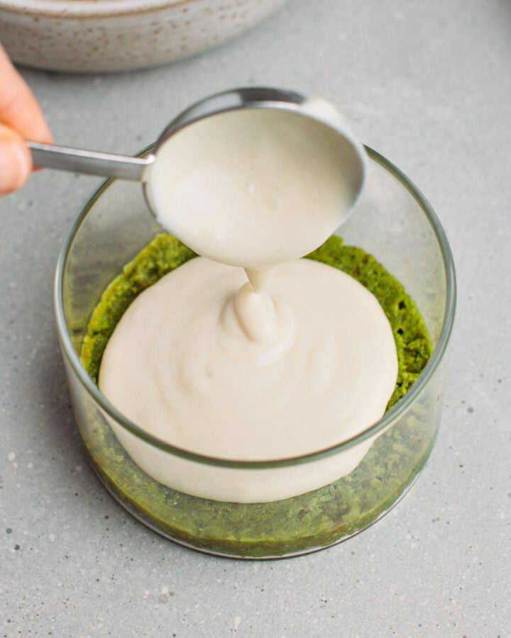 Pouring vanilla cream over a matcha sponge cake in a ramekin.
