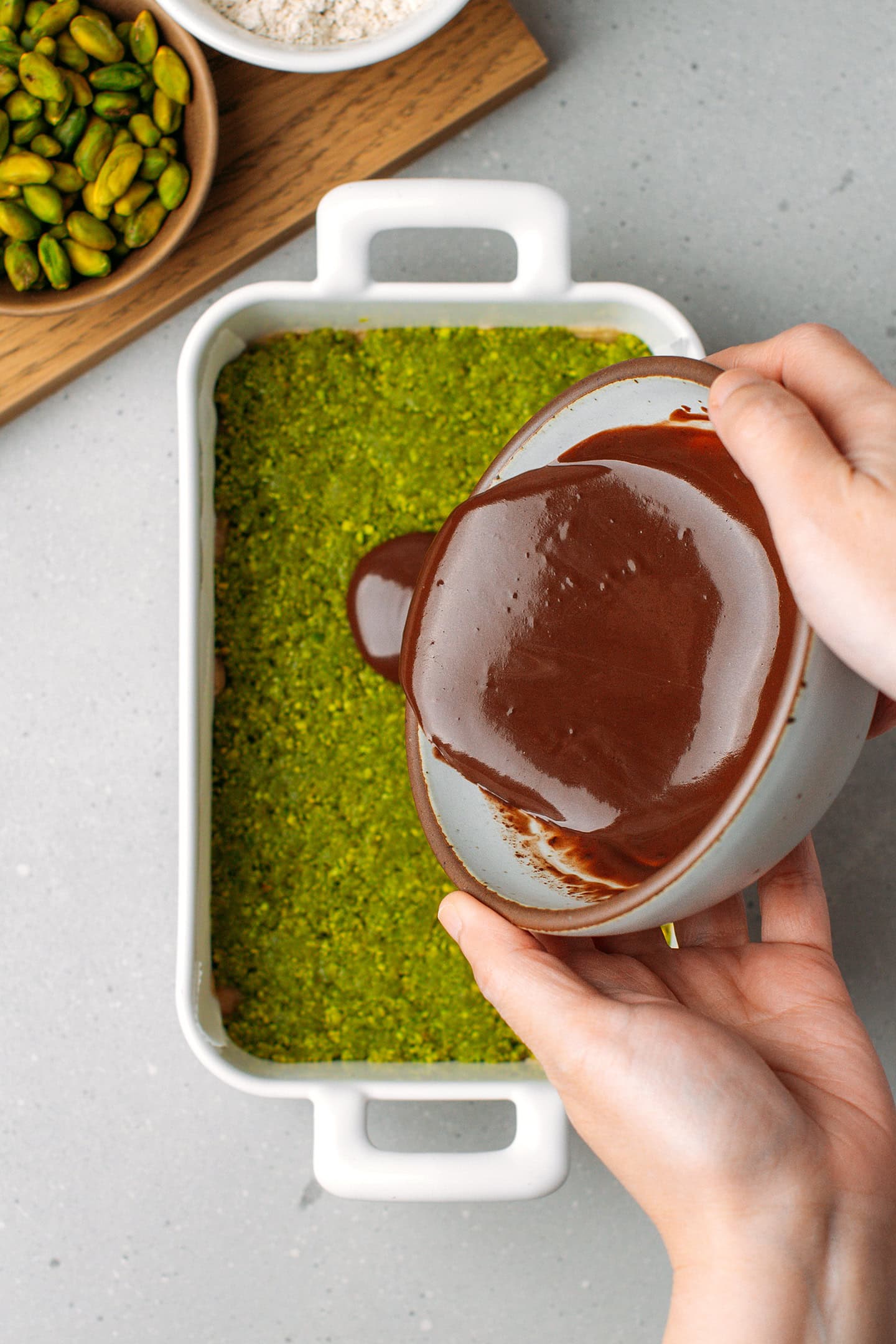 Pouring chocolate ganache over a layer of pistachio meal in a baking dish.