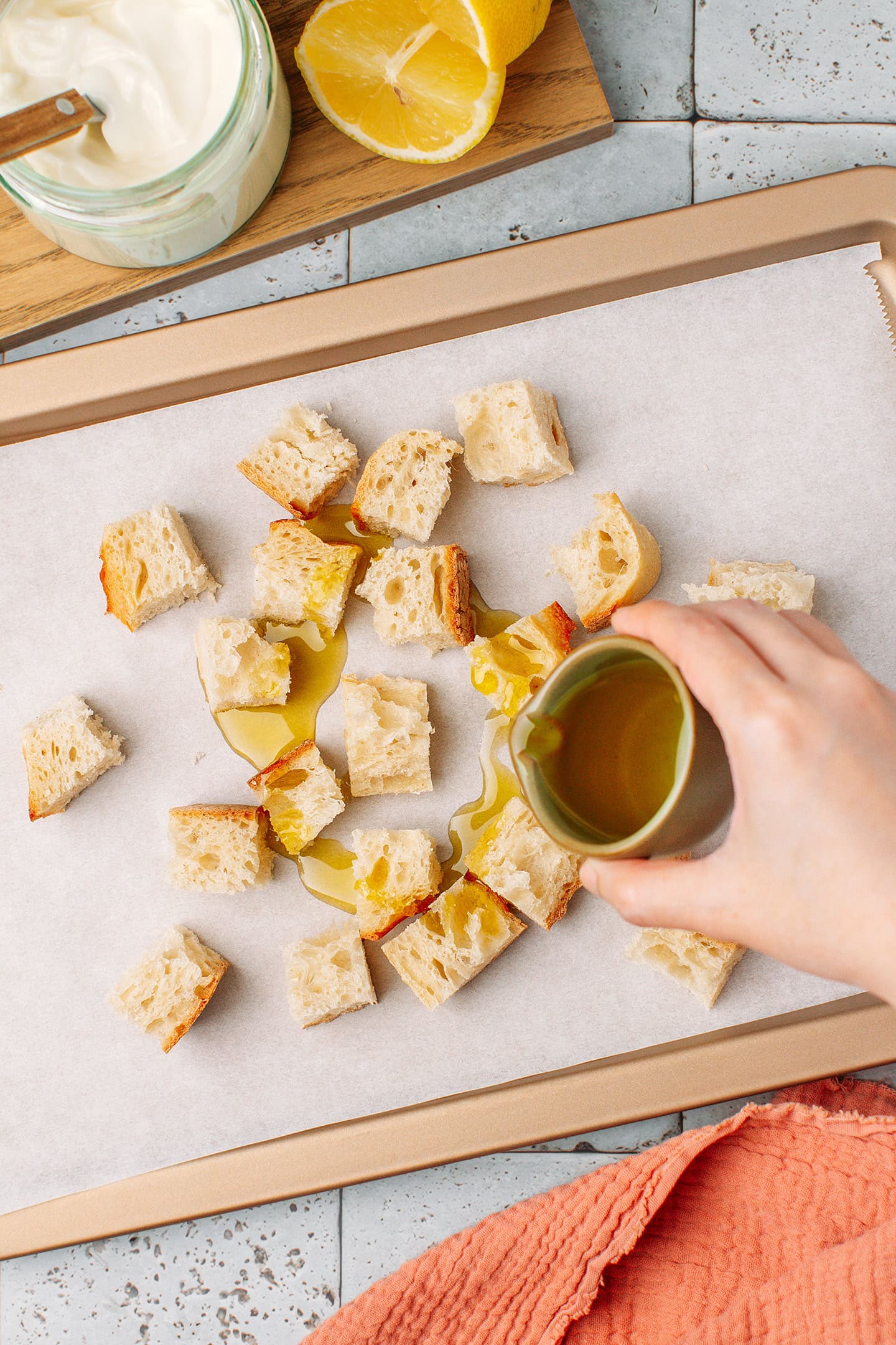 Pouring olive oil over diced bread on a baking sheet.