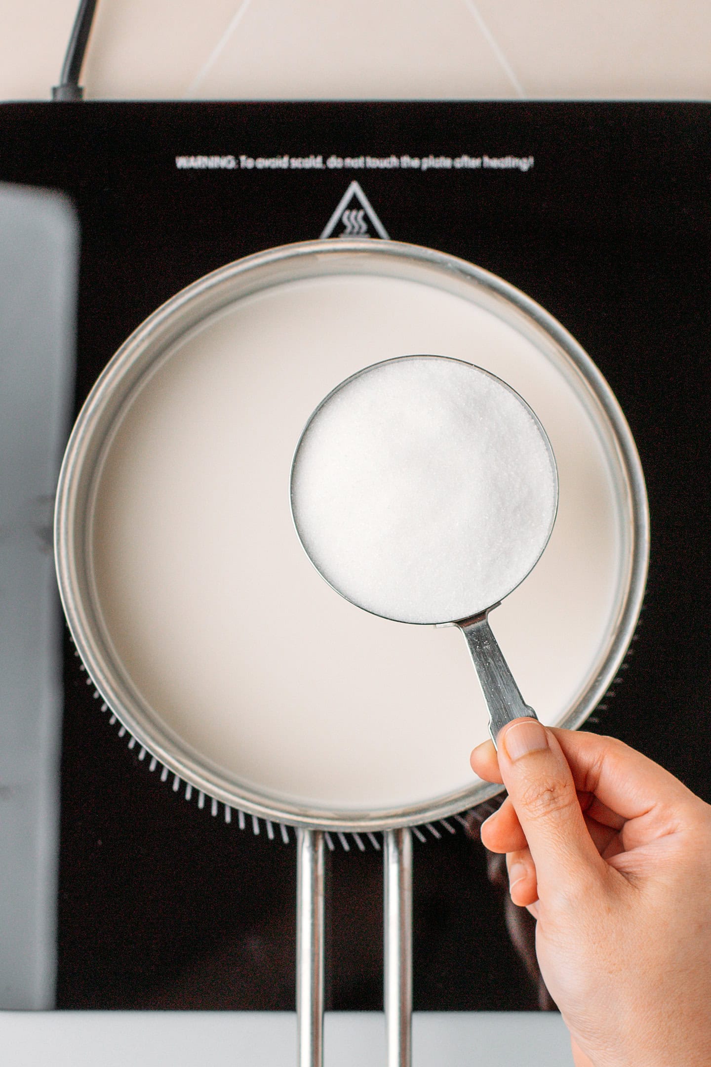 Holding half of a cup of sugar over a saucepan filled with almond milk.