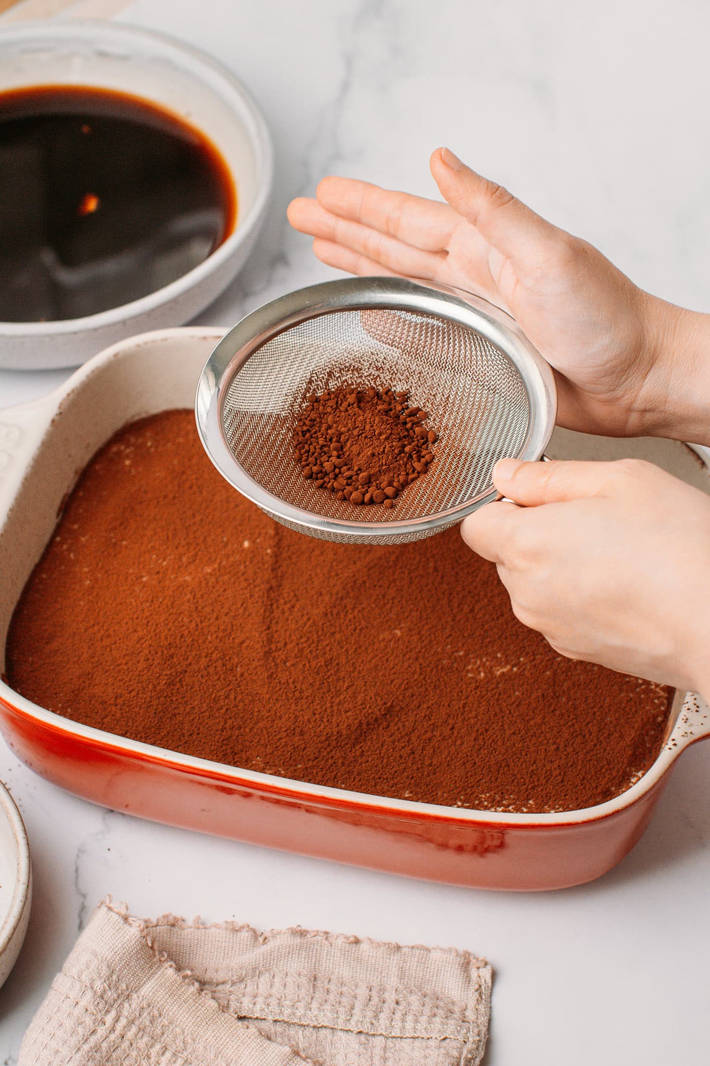 Dusting cocoa powder using a fine-mesh sieve over a baking dish.