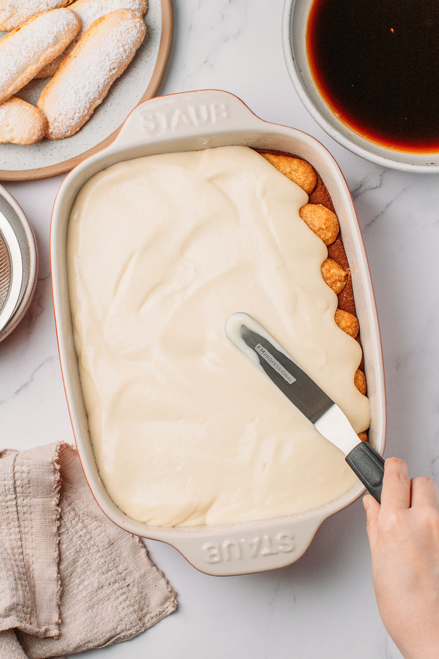 Spreading vanilla cream over ladyfingers inside a baking dish.