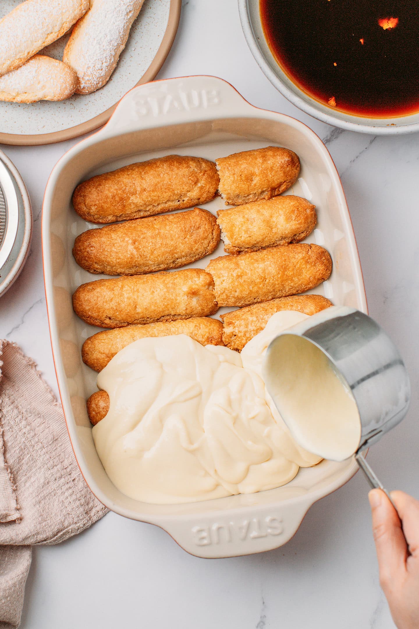 Pouring vanilla cream over ladyfingers in a baking dish.